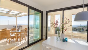 Una habitación moderna con un ventanal de vidrio que da a un balcón, una mesa blanca con un florero y muebles de mimbre.