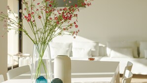 Una mesa de comedor blanca con un florero transparente que contiene flores rosas, un florero azul y un florero blanco con textura. Al fondo, hay un sofá blanco con almohadones y una pared blanca.