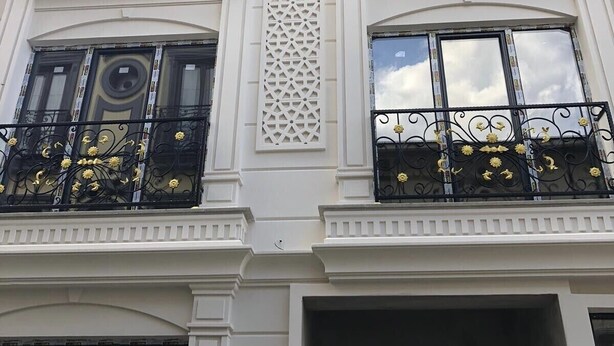 Un immeuble à plusieurs étages aux murs extérieurs blancs, doté de balcons en fer forgé noir ornés de motifs jaunes et de panneaux décoratifs en treillis.