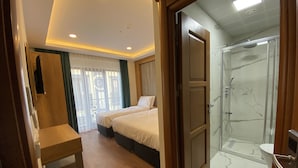 Une chambre d’hôtel avec deux lits, un bureau en bois, une douche aux carreaux de marbre et une vue sur l’extérieur par une fenêtre munie de rideaux.
