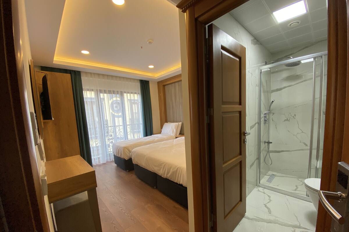 A hotel room with two beds, a wooden desk, a shower with marble tiles, and a view of the outdoors through a window with curtains.