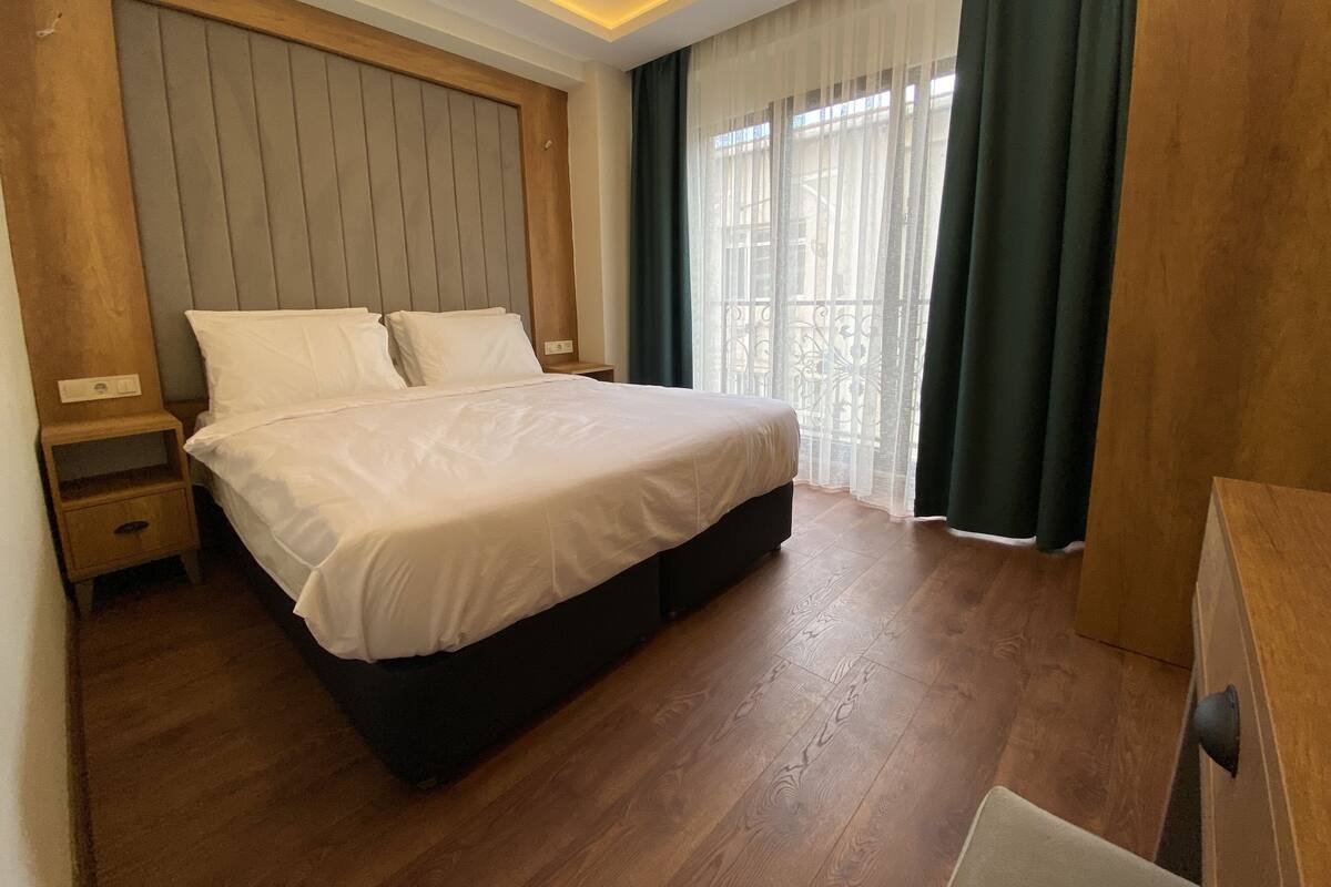 A modern hotel room with a large bed, wooden headboard, a bedside table, and a view of the outdoors through sheer curtains.