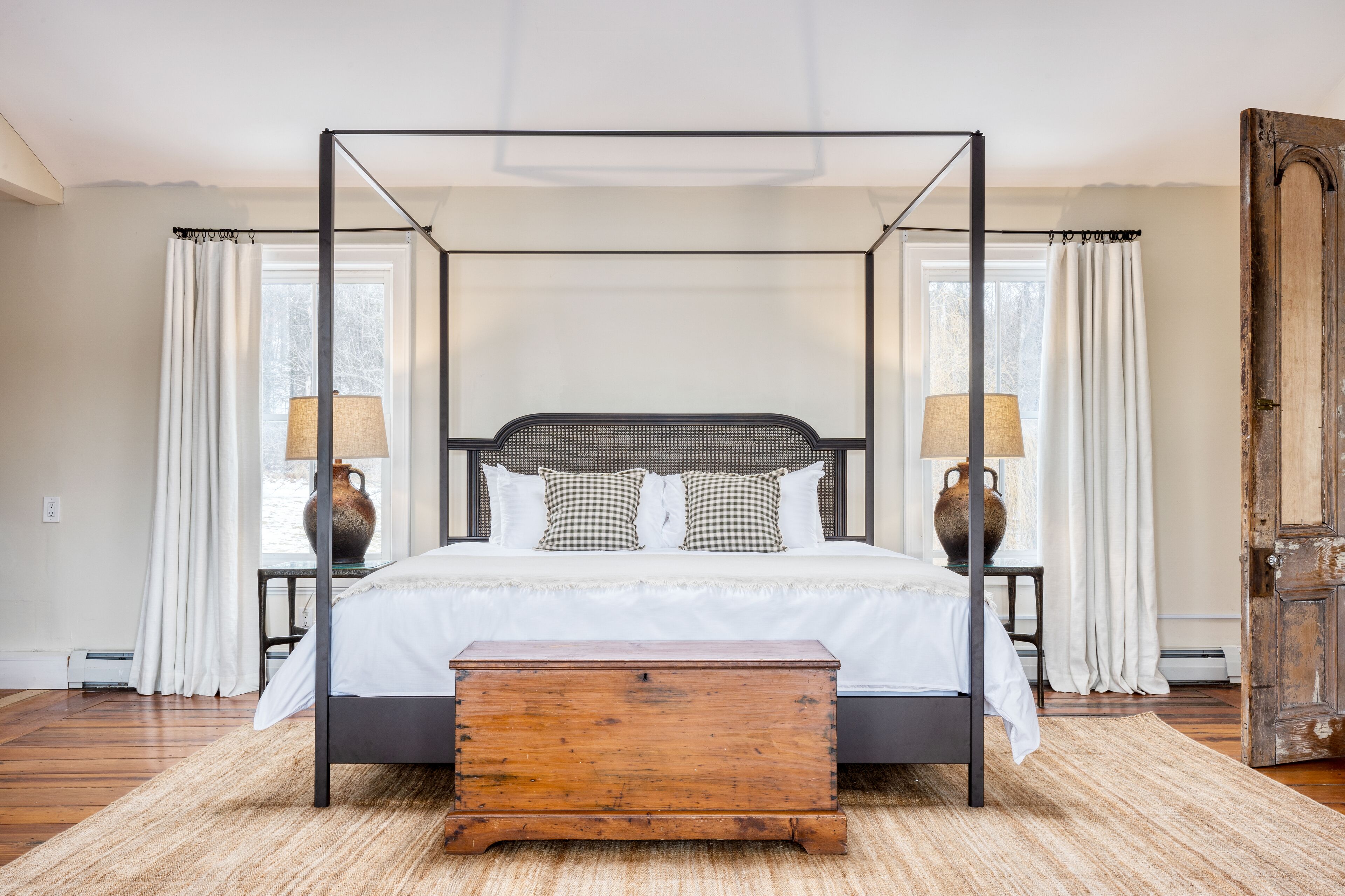 Una cama con dosel con cabecera de madera, ropa de cama blanca y almohadones cuadriculados. A cada lado de la cama hay una mesita de noche con una lámpara.