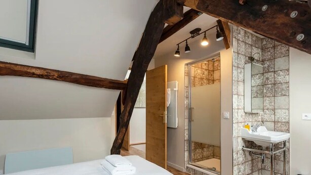 A bedroom with a bed, a wooden beam ceiling, a bathroom with a shower, and a sink.