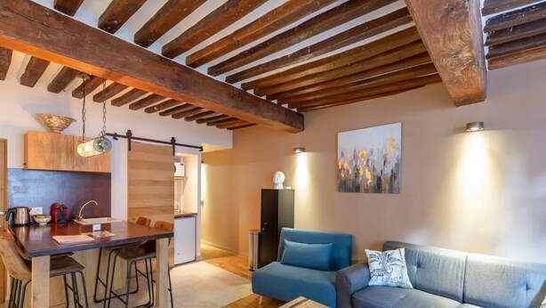 A cozy interior with exposed wooden beams, a dining area with stools, a sofa, and a painting on the wall.