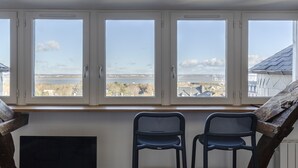 Ein Zimmer mit großem Fenster und Blick auf ein Gewässer sowie eine ferne Stadtlandschaft. Zwei schwarze Stühle und ein Flachbildfernseher stehen auf einem Holztisch.