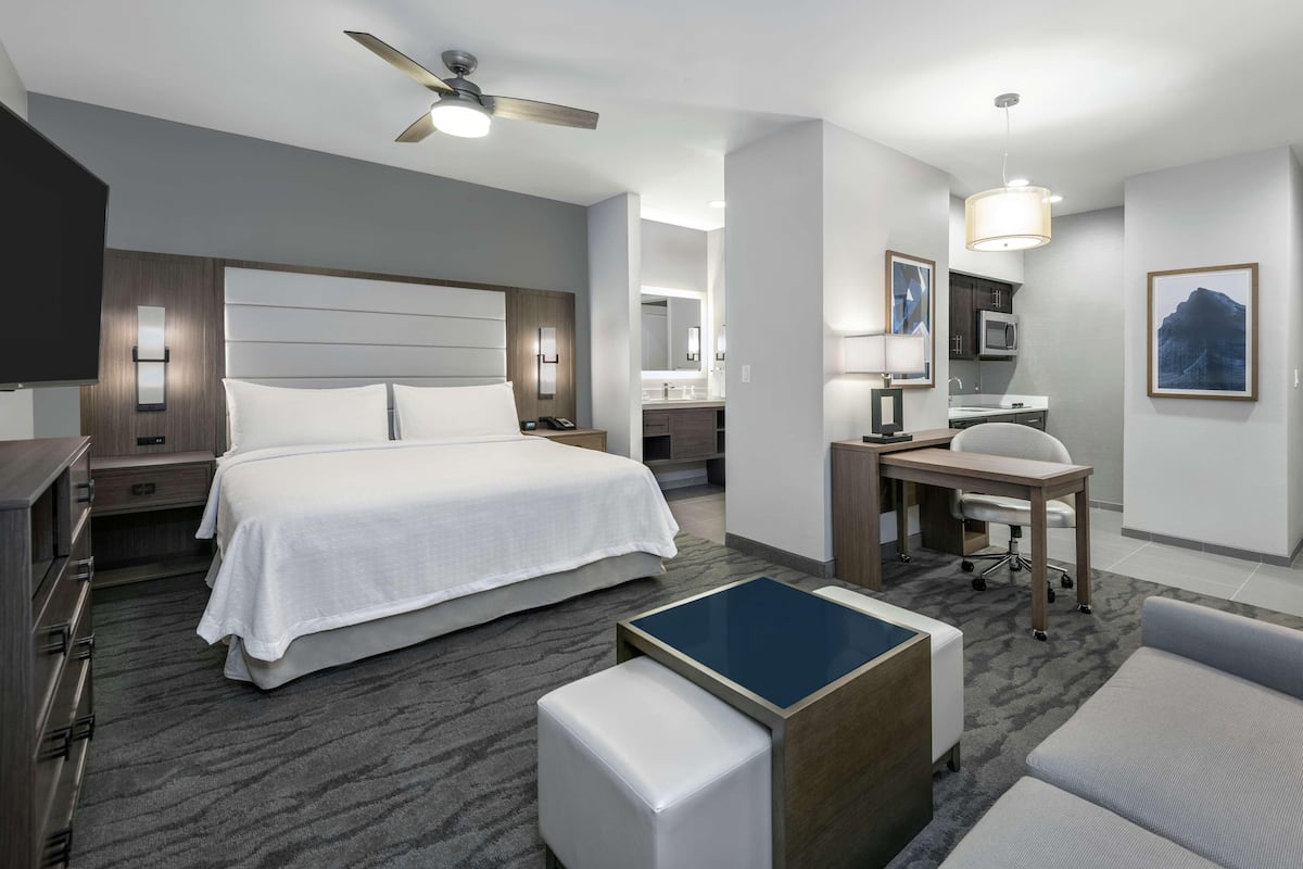 A modern hotel room with a large bed, a desk with a chair, a small table, a ceiling fan, and a bathroom visible in the background.