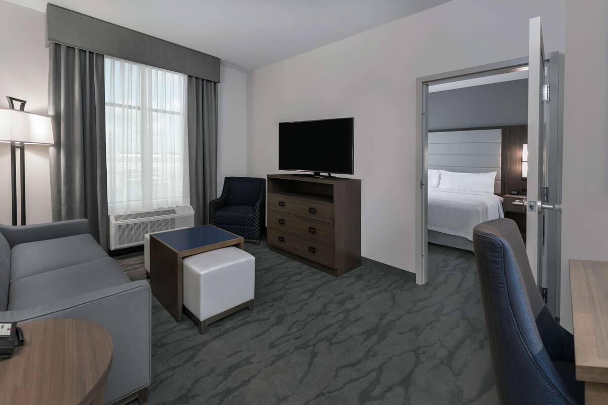 A hotel room with a sofa, a small table, a TV, a dresser, a bed, and a door leading to another room.