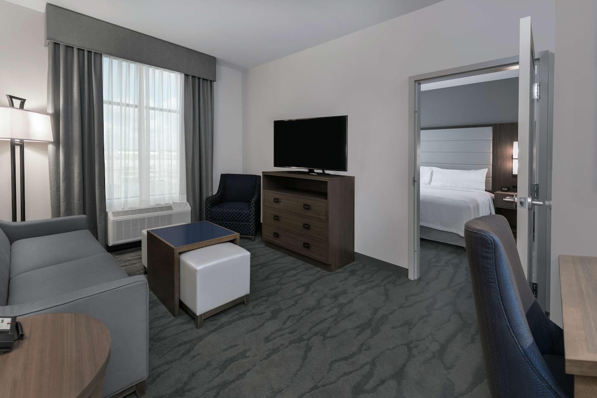 A hotel room with a sofa, a small table, a TV, a dresser, a bed, and a door leading to another room.