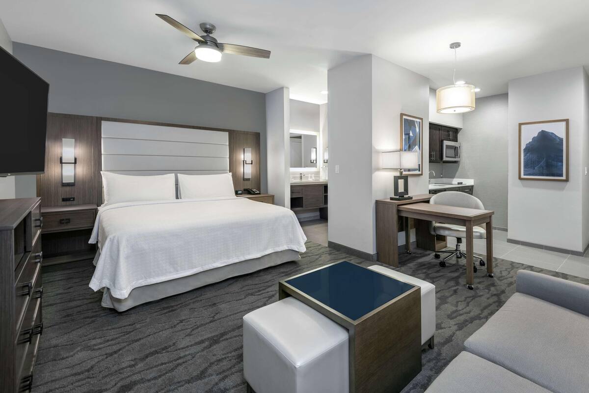 A modern hotel room with a large bed, a desk with a chair, a small table, a ceiling fan, and a bathroom visible in the background.
