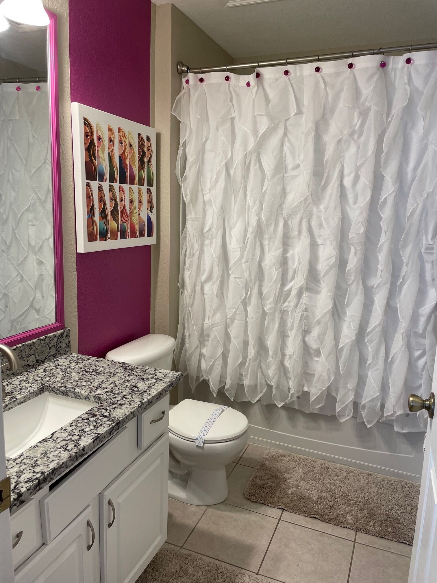 Une salle de bain avec des toilettes blanches, un lavabo sur un plan de travail en granit, un mur rose, un rideau de douche blanc et un tableau encadré représentant des femmes.