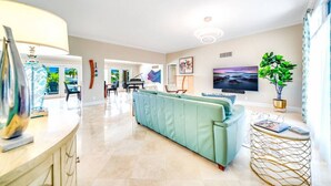 A spacious living room with a light blue sofa, a piano, a television, and a large window with a view of greenery. - EXQUISITE HEATED POOL+WATERFRONT! 1 MILE TO BEACH! (Pompano Beach)