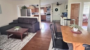 A living room with a sofa, coffee table, and dining area with chairs and a table. A kitchen with appliances and a door with a wreath is visible in the background. - The Perfect Private Escape (Lorraine)