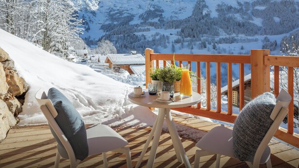 Een houten terras met twee witte stoelen en een tafel, met uitzicht op een besneeuwd berglandschap met huizen.