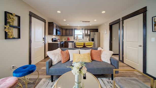 A modern living room with a grey sofa, a round coffee table, and a blue chair. A kitchen is visible in the background with a refrigerator, microwave, and yellow chairs. - Lovely 3 bedroom Apartment in Bozeman! Unit #C  (Bozeman)