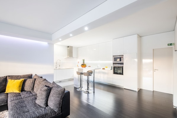 A modern living room with a sofa and a coffee table, a kitchen with white cabinets and appliances, and a wooden floor.