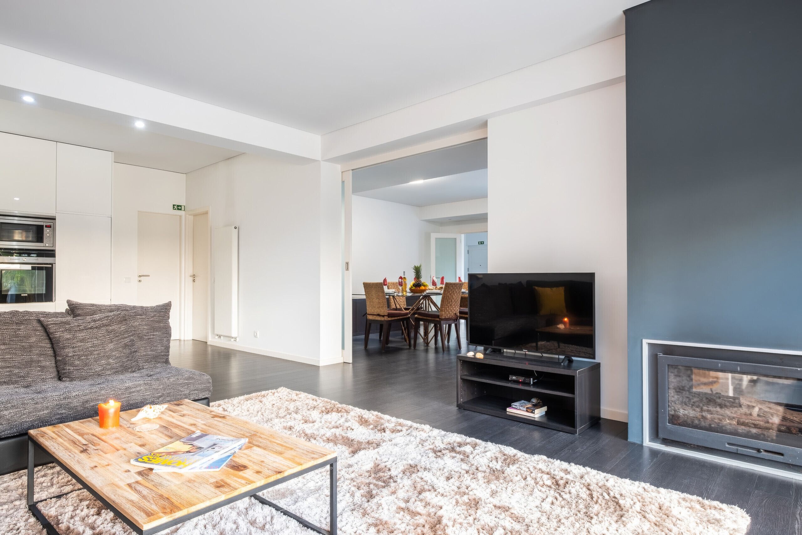 A modern living room with a flat-screen TV, a fireplace, a dining area, and a kitchen with built-in appliances.
