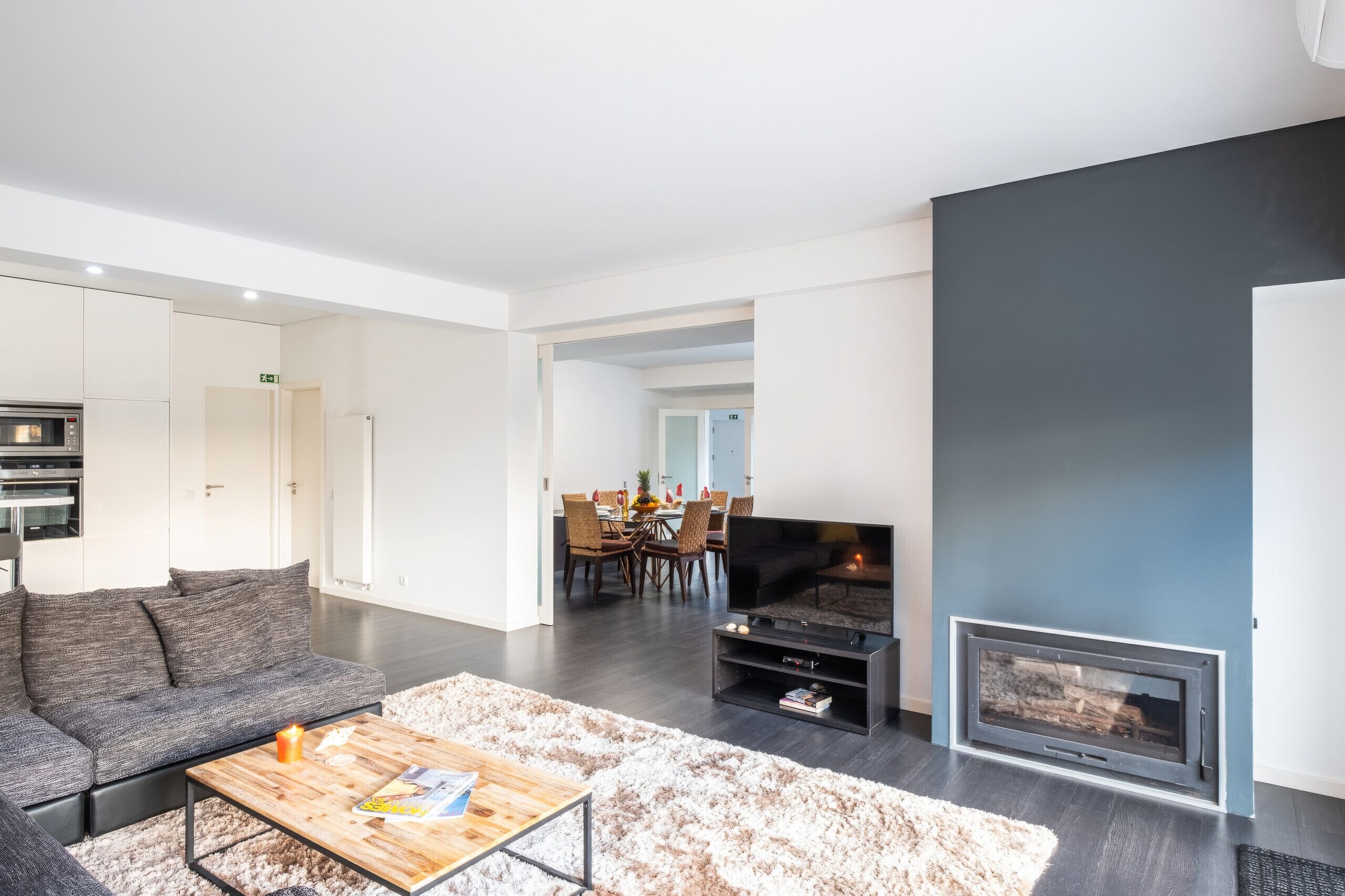 A modern living room with a sofa, coffee table, fireplace, and a dining area in the background.