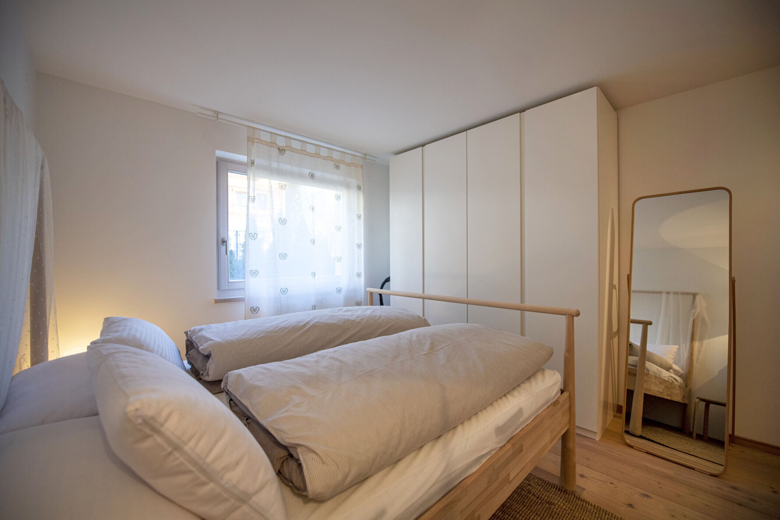 A bedroom with two beds, a window with heart-patterned curtains, a mirror, and a wardrobe.