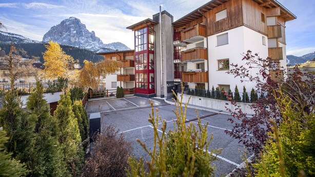 Ein modernes, mehrstöckiges Gebäude mit einem zentralen Glasaufzugsschacht, umgeben von einem Parkplatz und üppigem Grün.