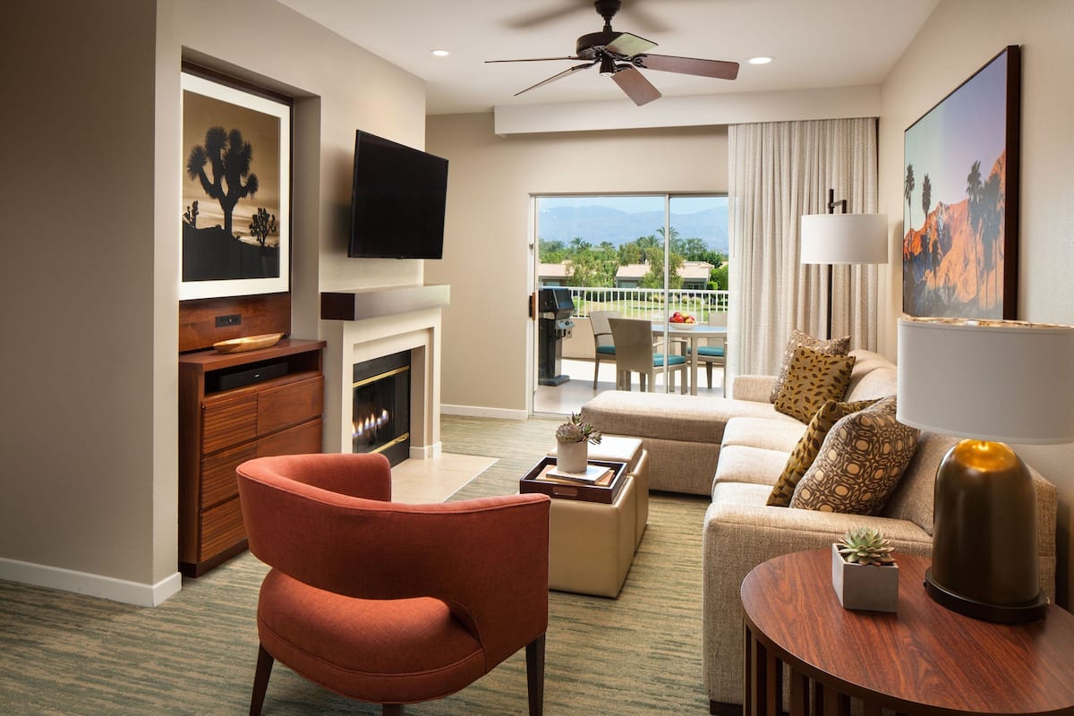 A modern hotel room with a fireplace, a flat-screen TV, a ceiling fan, a balcony with a view of mountains, and a dining area.