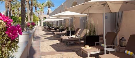 Un espacio lounge al aire libre, iluminado por el sol, con varias opciones de asientos, incluyendo sillones y sofás, todo bajo grandes sombrillas.
