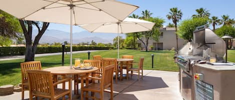 An outdoor patio with wooden tables and chairs, a large umbrella, and a built-in grill.