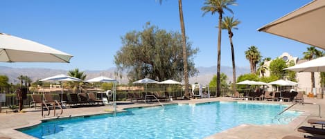 A swimming pool with lounge chairs and umbrellas, surrounded by palm trees and mountains in the background.