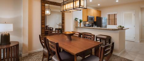 A dining area with a wooden table and chairs, a central light fixture, and a kitchen in the background.