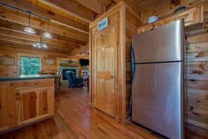 A wooden cabin interior with a kitchen area, including wooden cabinets, a refrigerator, and a stone fireplace. - Maple Hill Cabin | Pet-Friendly Lake Lure Cabin with Views, Fireplaces & Riverbend Amenities (Lake Lure)