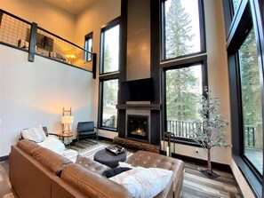 Una sala de estar moderna con chimenea, ventanas amplias, una escalera y vistas a árboles.