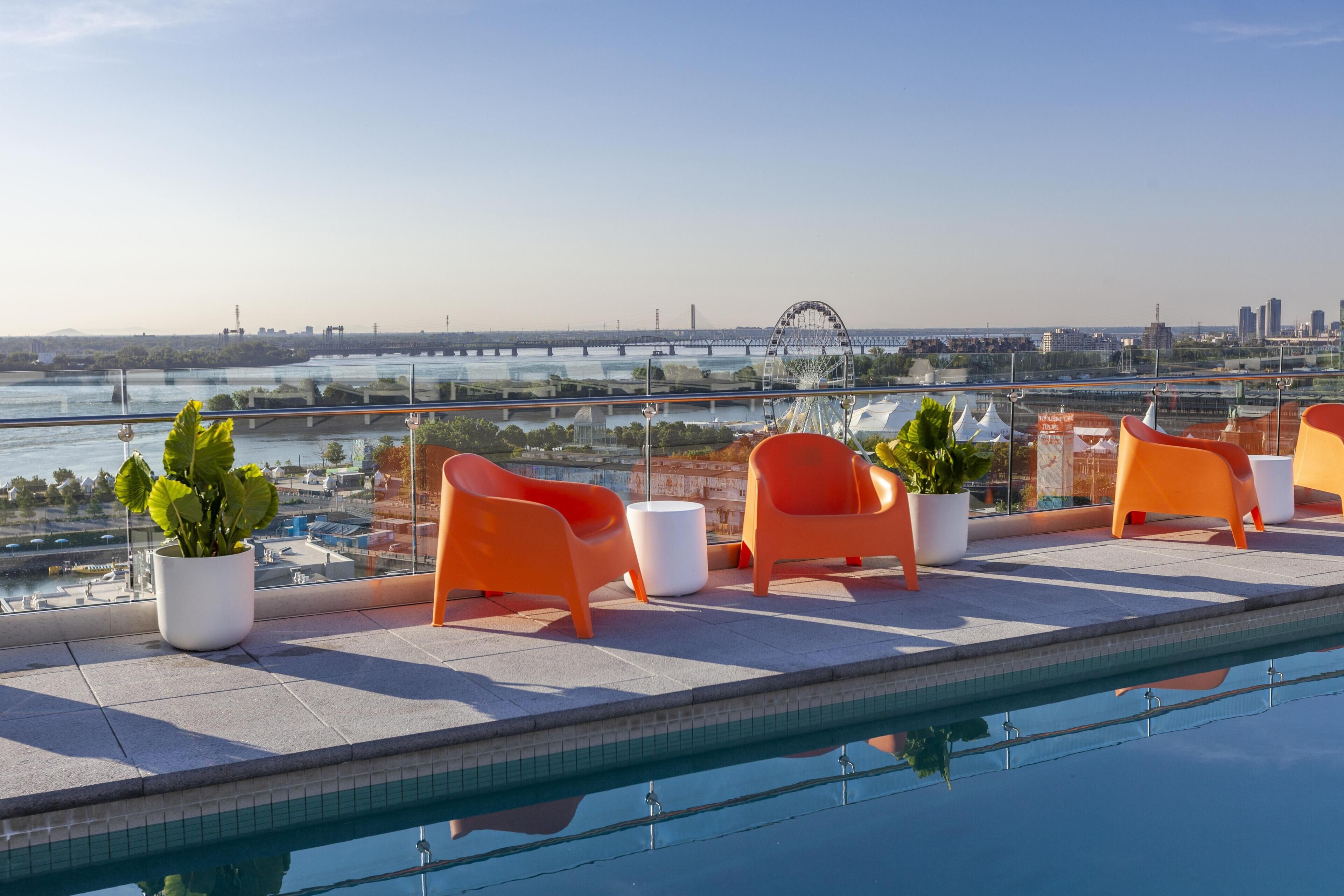 Piscina en la azotea con sillas anaranjadas y plantas en macetas, con vistas a un paisaje urbano que incluye un puente y edificios.