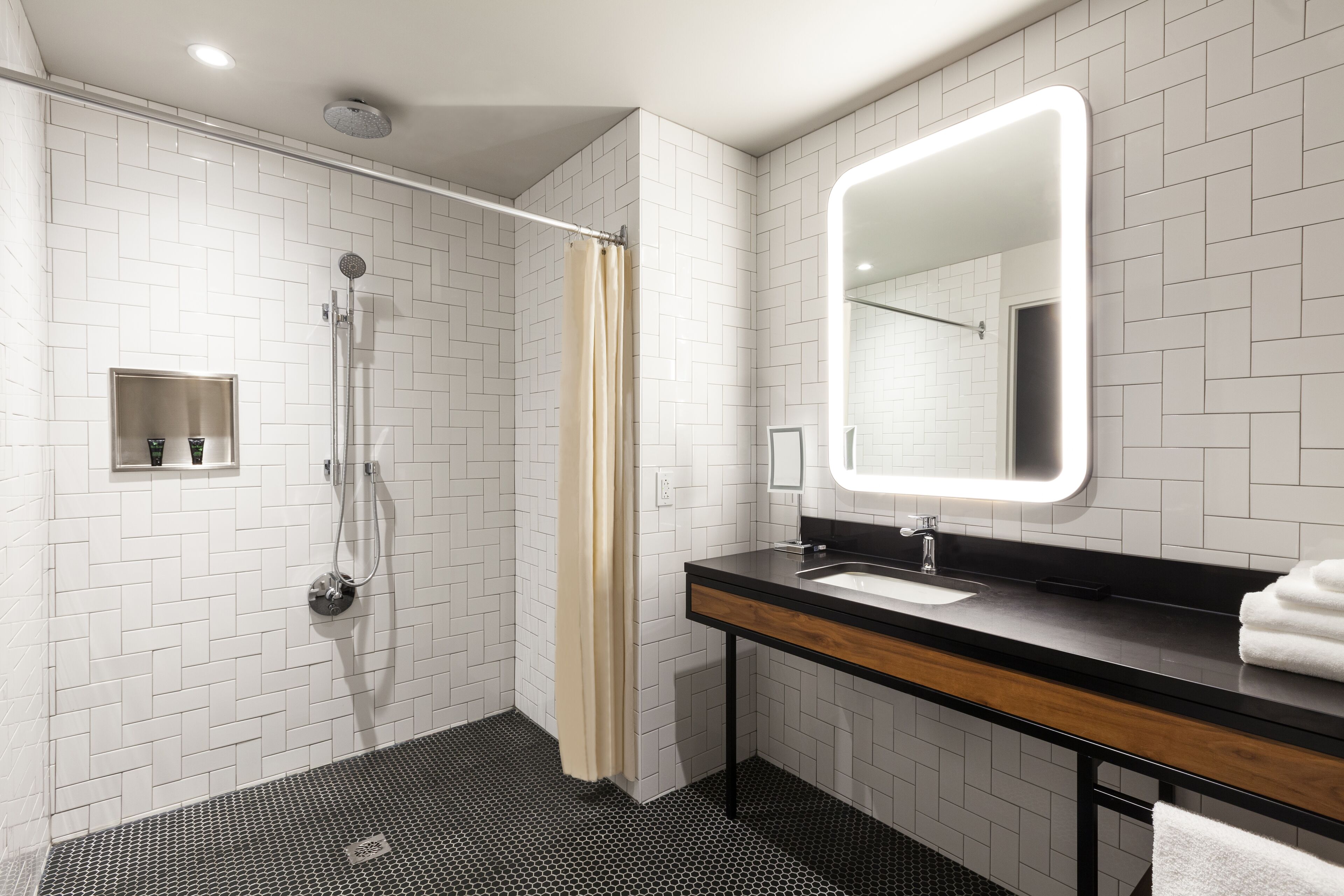 Une salle de bain moderne avec des murs recouverts de carreaux blancs, un sol à carreaux noirs et blancs, un meuble-lavabo en bois, un grand miroir et une douche avec un rideau beige.