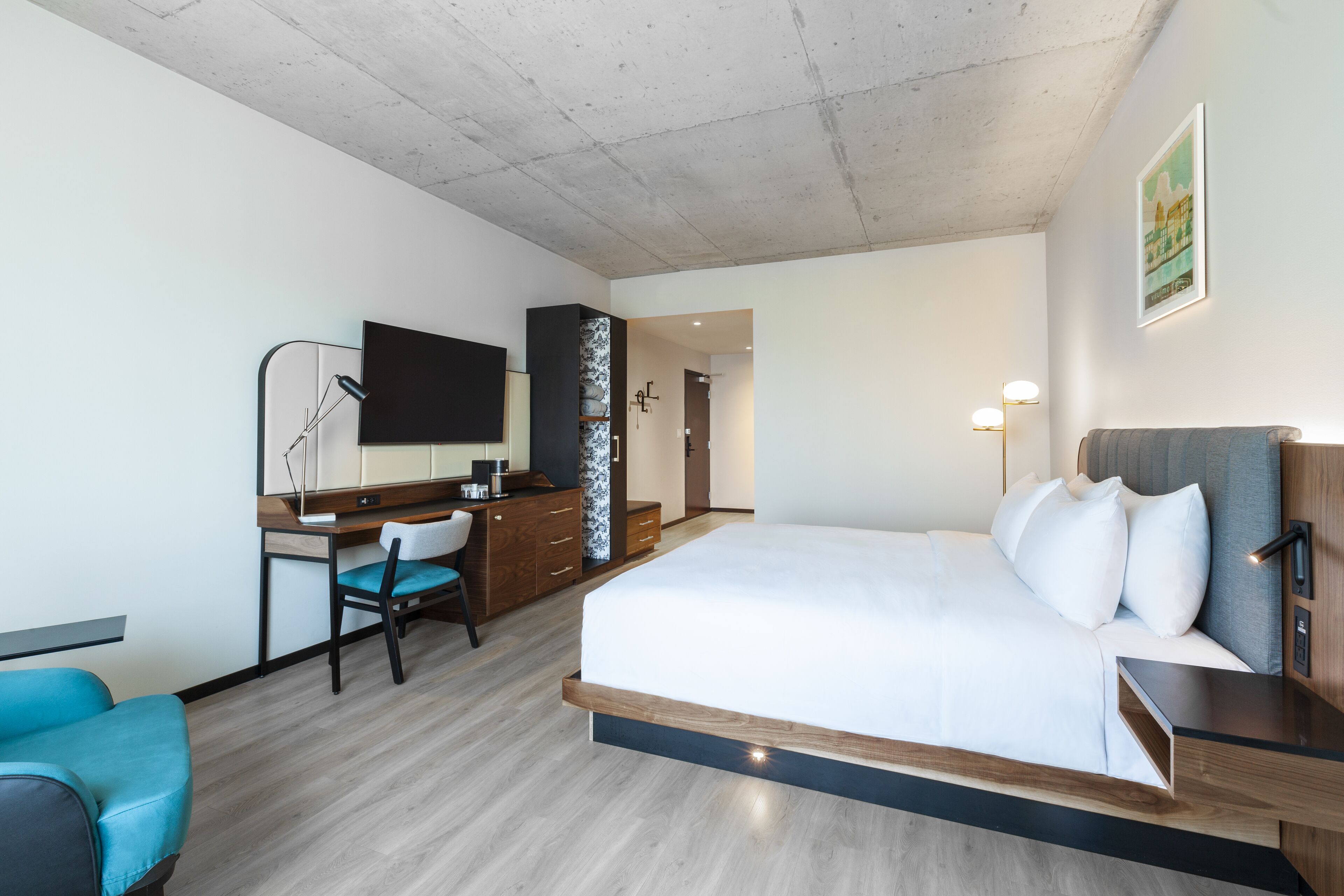 A modern hotel room with a large bed, a desk with a chair, a flat-screen TV, and a small kitchenette.