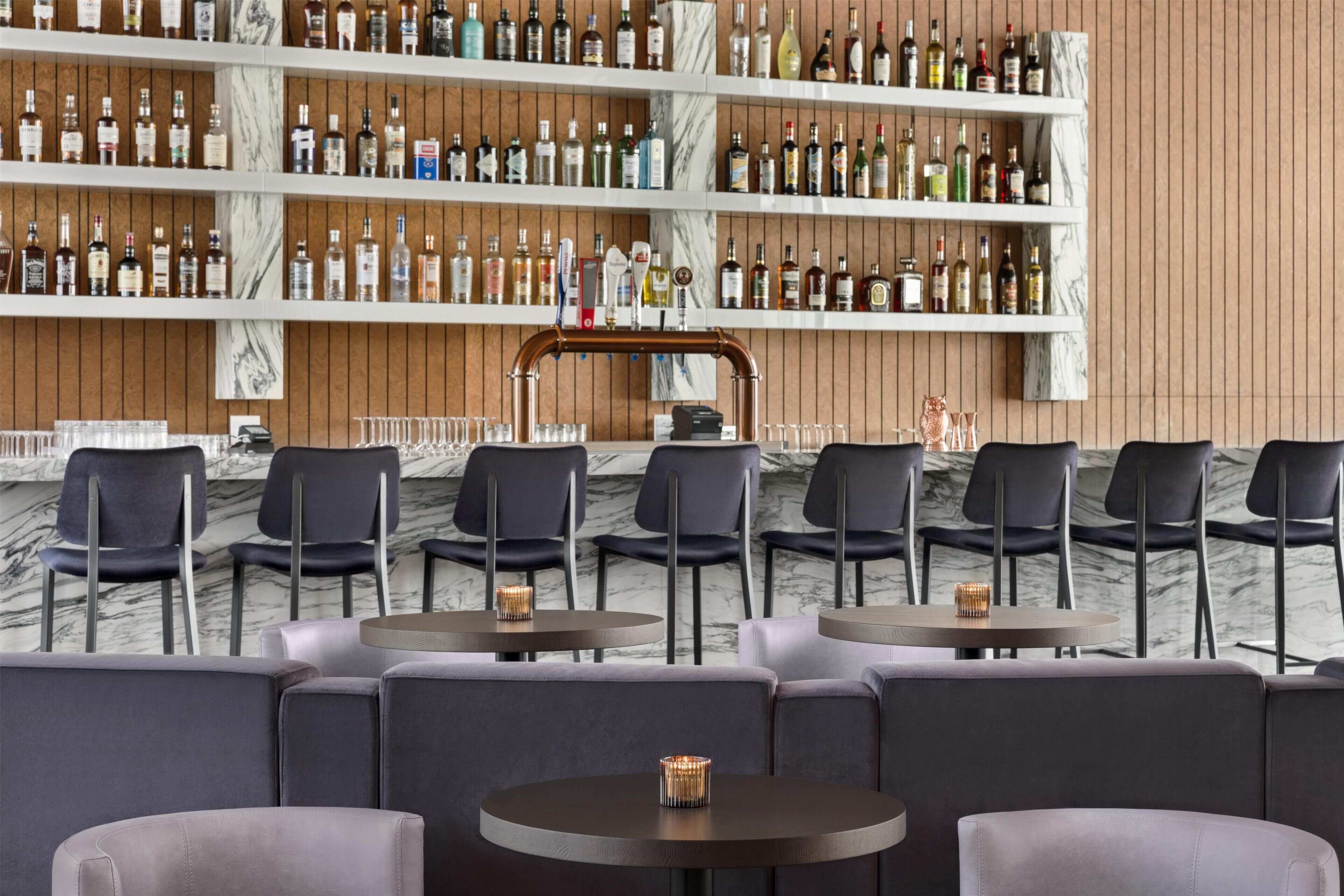 A bar area with multiple shelves of liquor bottles, round tables with chairs, and a marble wall.