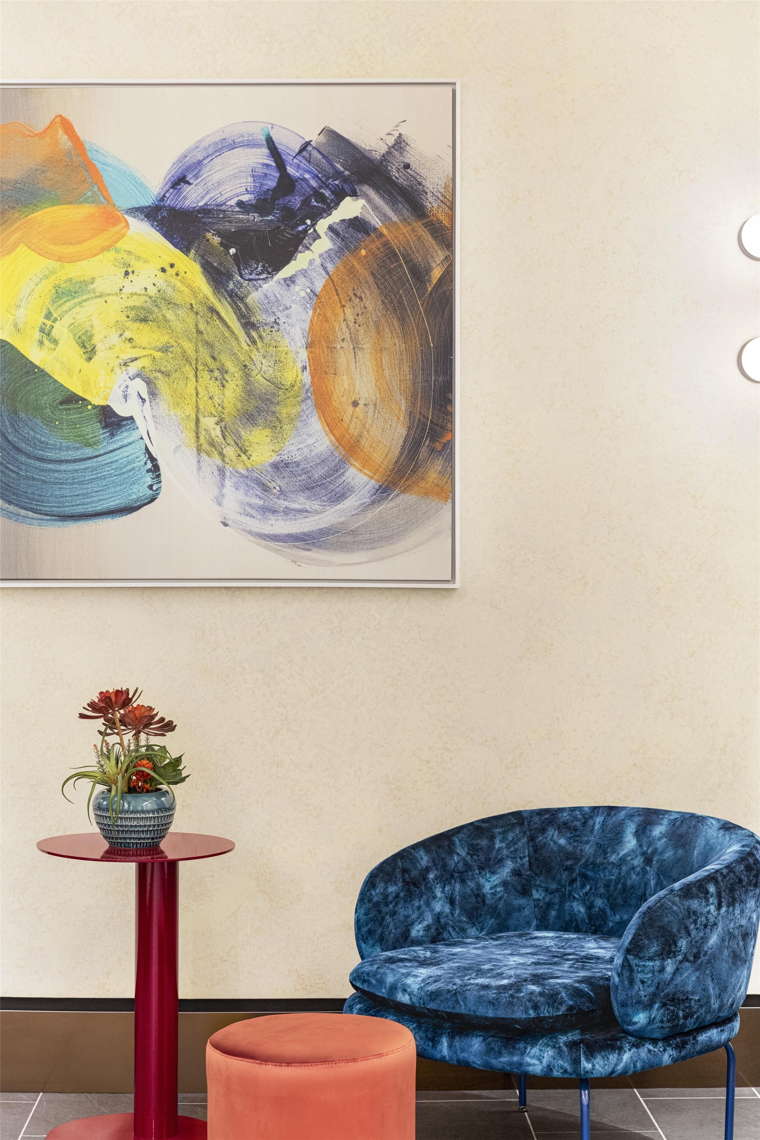 A room with a blue velvet chair, a red table with a vase, and a large abstract painting on the wall.