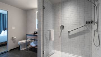 A modern bathroom with a glass shower enclosure, a toilet, a sink with a mirror, and a bed in the adjacent room.