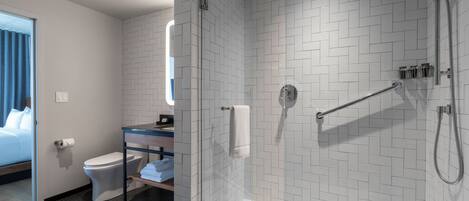 Un baño moderno con cabina de ducha de vidrio, inodoro, lavabo con espejo y una cama en la habitación contigua.