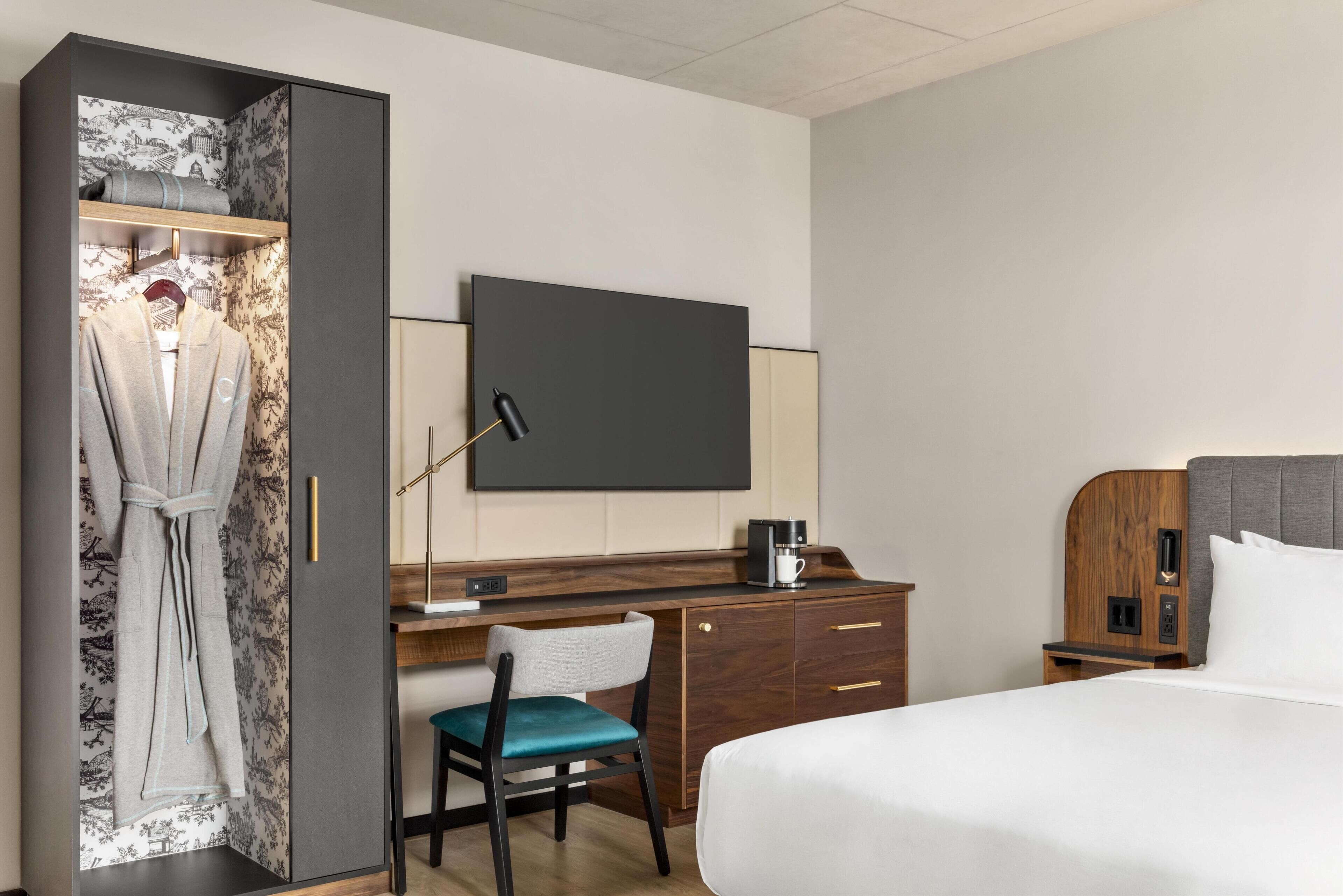 Habitación de hotel con cama, escritorio con silla, armario, televisor de pantalla plana y cafetera.