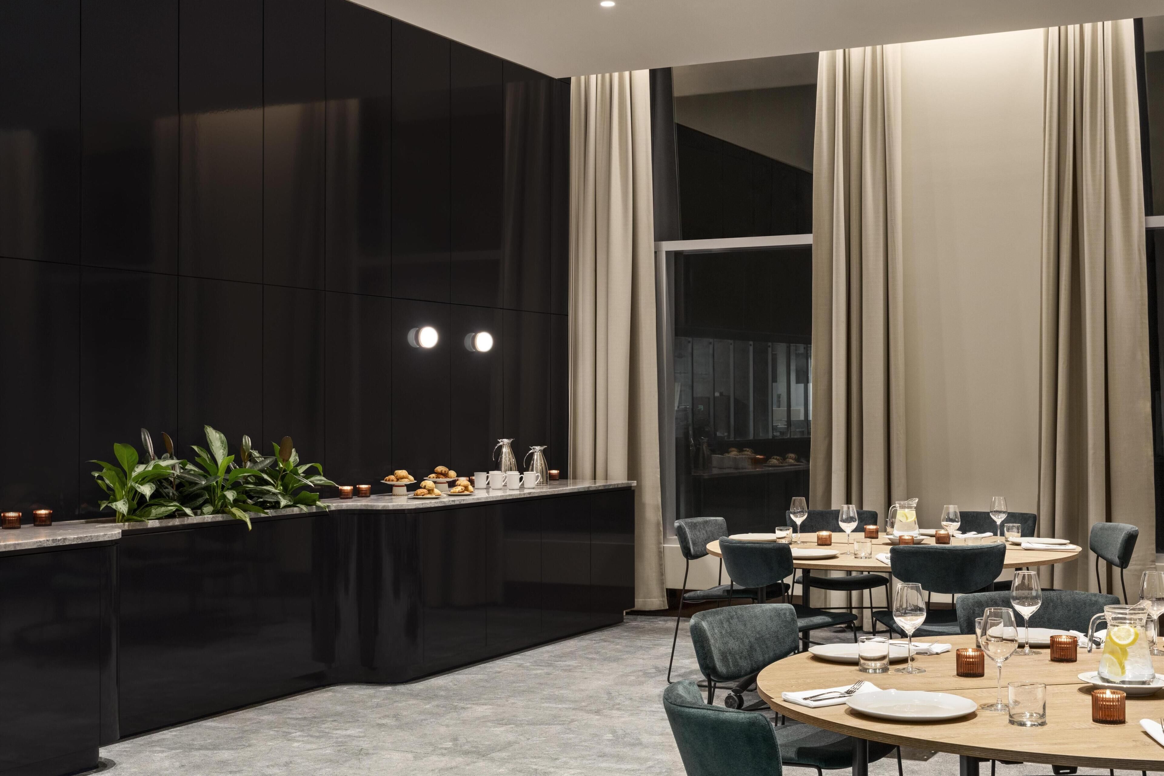 Un comedor moderno con una barra negra, plantas verdes y una mesa preparada para cuatro personas, con vasos y cubiertos.