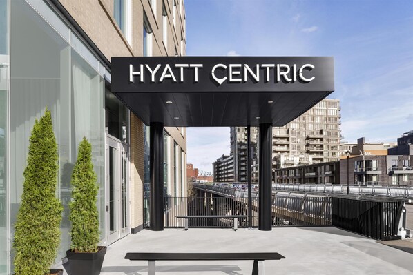 The exterior of the Hyatt Centric hotel with a clear sky, a modern entrance, and a view of the surrounding urban area.