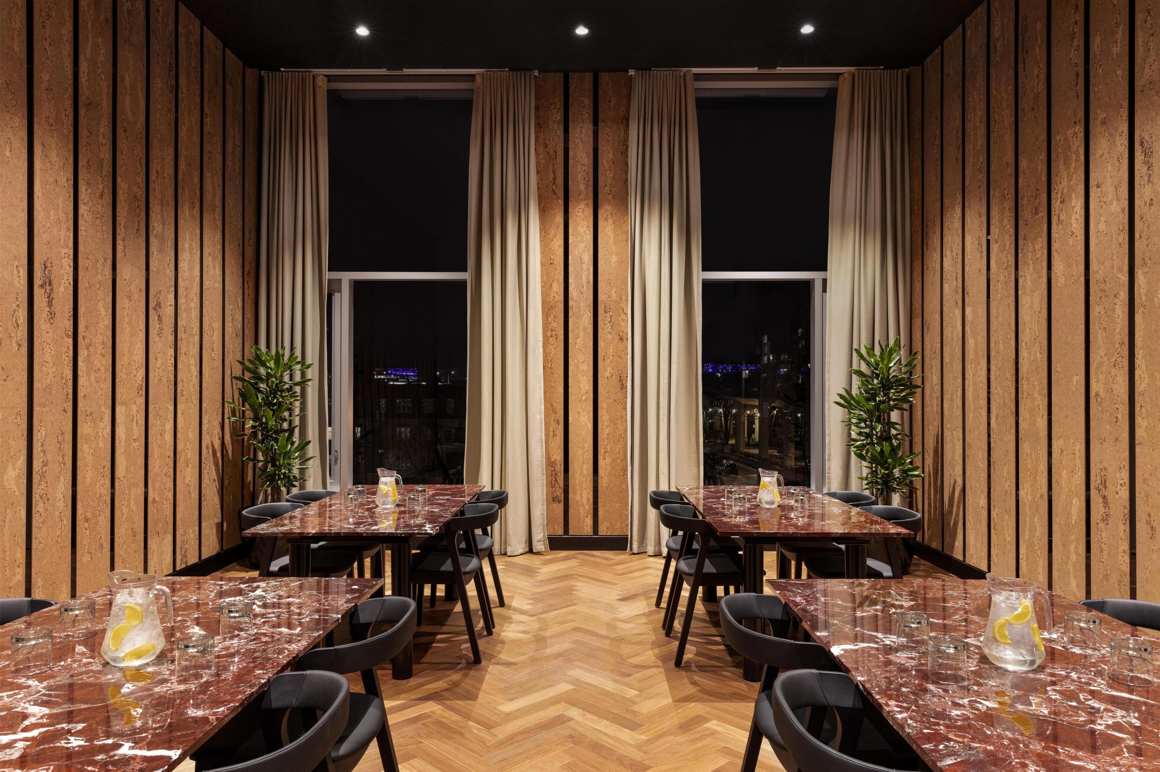 A restaurant with wooden panel walls, large windows with curtains, and a herringbone floor. Tables are set with glasses and pitchers.