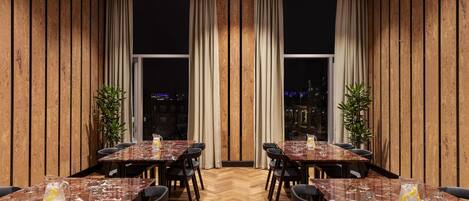 A restaurant with wooden panel walls, large windows with curtains, and a herringbone floor. Tables are set with glasses and pitchers.