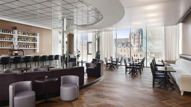 En modern hotellobby med en bar, sittgrupper och stora fönster med utsikt över staden.