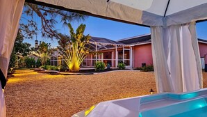 Un área de piscina con un porche cubierto, un jardín ajardinado con palmeras y un edificio de fachada rosa.