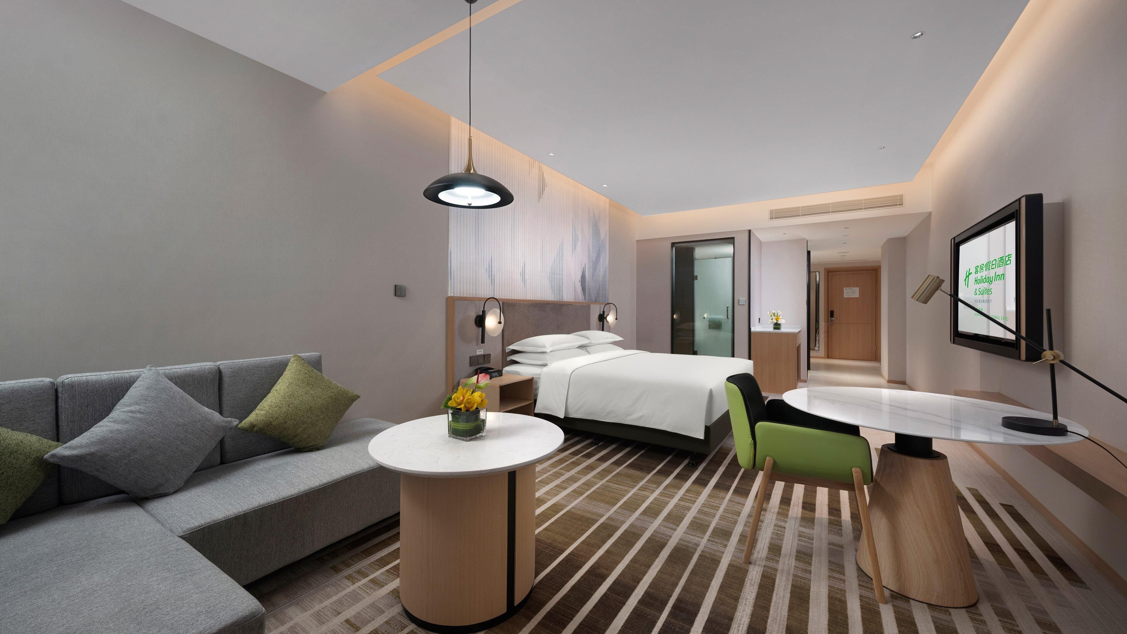 Una habitación de hotel moderna con un sofá gris, una cama con sábanas blancas, una mesita redonda con un florero y un escritorio con una silla.