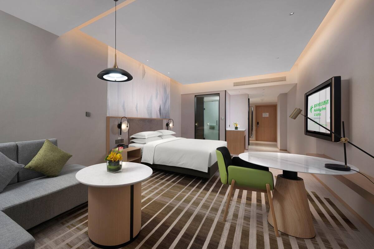 Une chambre d’hôtel moderne avec un canapé gris, un lit avec du linge de maison blanc, une petite table ronde ornée d’un vase de fleurs et un bureau avec une chaise.
