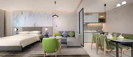 A modern hotel room with a bed, a sofa, a dining table with chairs, a small kitchen area, and a bathroom.