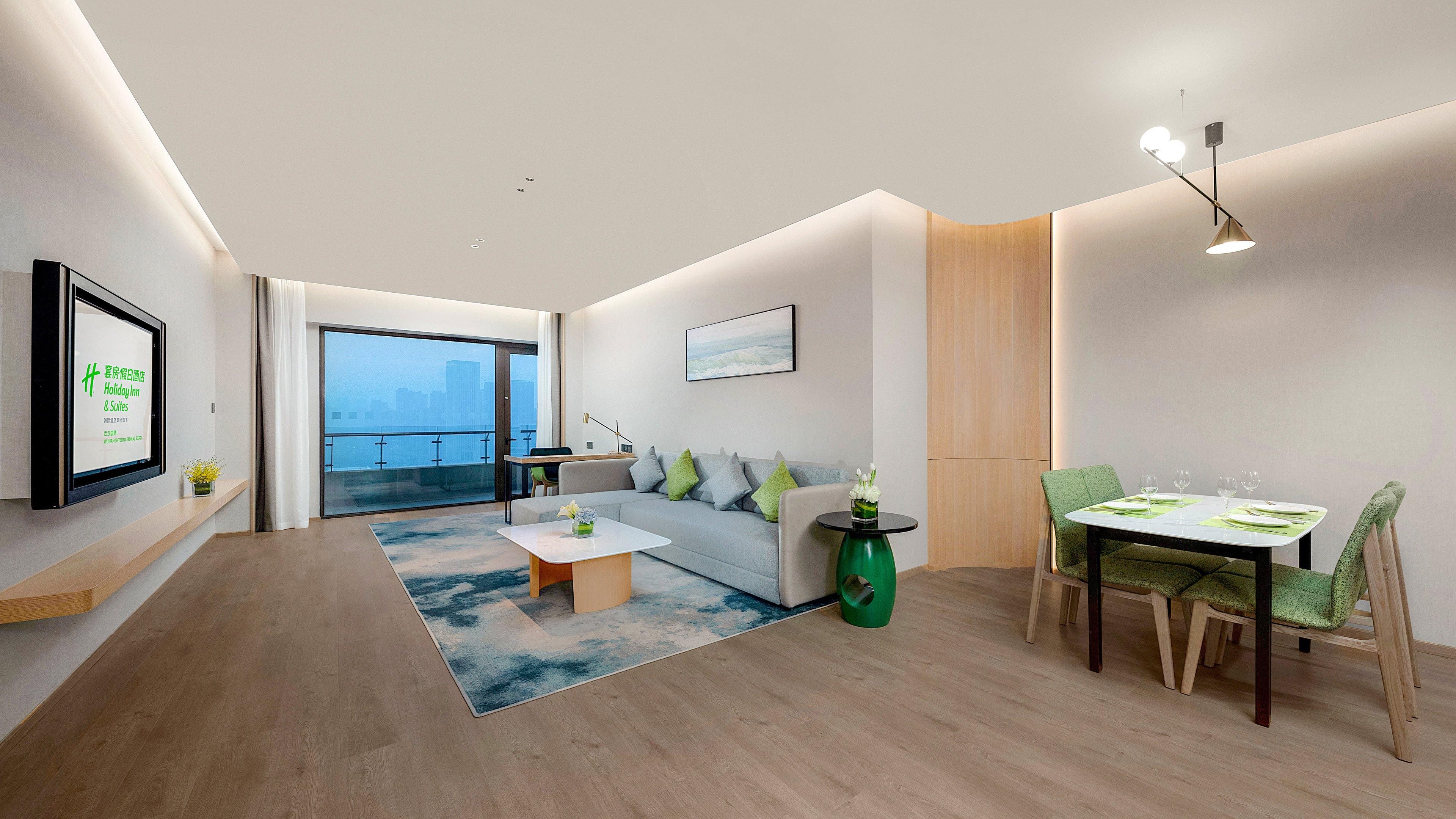 Quarto de hotel moderno com sofá, mesa de jantar e uma grande janela com vista para a cidade.