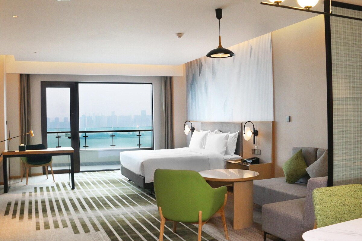 Ein Hotelzimmer mit Bett, Schreibtisch, Sessel, Sofa, Fernseher und Blick auf die Stadt.